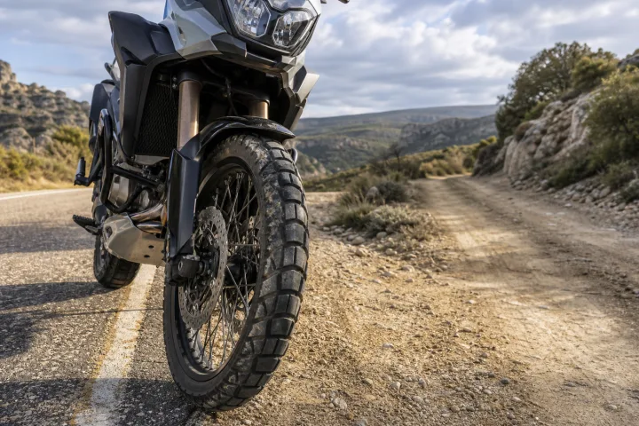 Moto trail estacionada sobre asfalto junto a un camino de tierra, con el neumático delantero en primer plano.